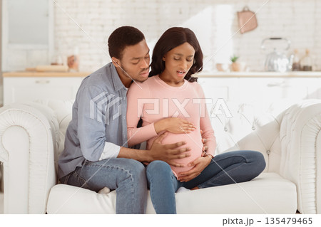 African American pregnant woman suffering from pain, having birth contractions while worried husband helping her at home. Beautiful expectant lady in labor with her boyfriend African American pregnant woman suffering from pain, having birth contractions while worried husband helping her at home. Beautiful expectant lady in labor with her boyfriend 135479465