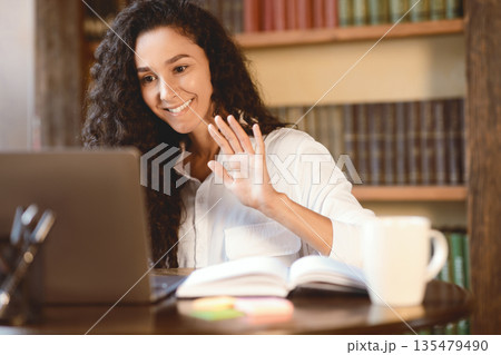 Virtual Video Conference. Portrait of smiling young woman making online call on laptop, waving with hand to webcam. Communication Concept. Woman chatting using computer, sitting at desk Virtual Video Conference. Portrait of smiling young woman making online call on laptop, waving with hand to webcam. Communication Concept. Woman chatting using computer, sitting at desk 135479490
