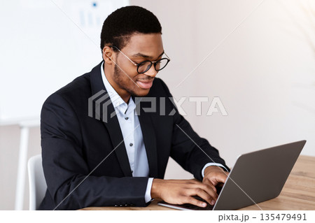 Smiling young black manager in formal suit working with laptop, wearing glasses, office interior, copy space. Successful millennial african american businessman working on business project 135479491