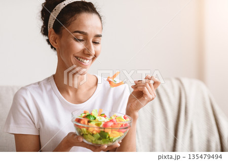 Closeup of smiling beautiful young woman holding bowl of healthy salad and fork, sitting on sofa at home, copy space. Healthy young brunette woman enjoying vegetables for lunch. Diet concept Closeup of smiling beautiful young woman holding bowl of healthy salad and fork, sitting on sofa at home, copy space. Healthy young brunette woman enjoying vegetables for lunch. Diet concept 135479494