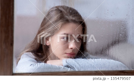 Unhappy young lady looking out window with drops of rain, feeling sad or melancholic. Emotional woman suffering from seasonal depression or loneliness. Mental health concept 135479507