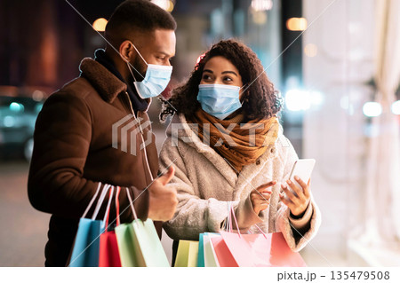 Retail, Technology And Pandemic Concept. Portrait of african american couple wearing medical masks, using smartphone, lady pointing at screen and showing app to her husband, holding shopping bags 135479508