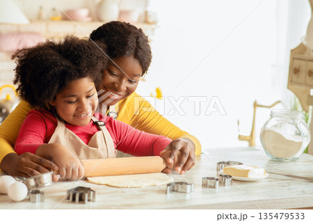 Happy black mom and her daughter rolling up dough together on table in kitchen, having fun while preparring homemade pastry, loving african american family enjoying spending time together at home 135479533