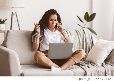 Angry woman freelancer looking at laptop screen and talking on phone, living room interior, copy space. Emotional young brunette lady having problems with her job, sitting on couch at home 135479552