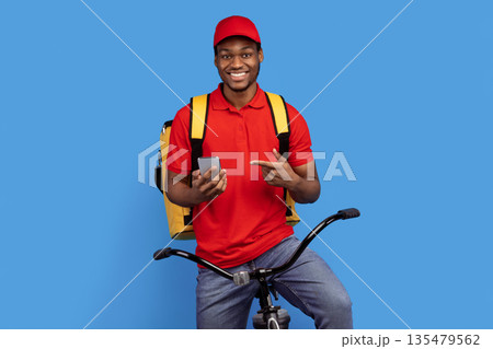Courier Service Application. Smiling black delivery guy using smartphone and pointing at it, wearing red uniform, cap and thermo bag, riding bicycle, isolated on blue studio background 135479562