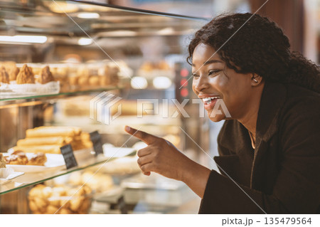Delicious dessert in store or candy shop and discounts for sweets. Focus on smiling young african american woman points her finger at cake in showcase and want to buy, close up, copy space Delicious dessert in store or candy shop and discounts for sweets. Focus on smiling young african american woman points her finger at cake in showcase and want to buy, close up, copy space 135479564