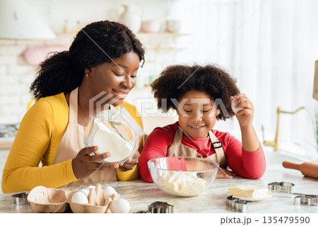 Cheerful black mother and little daughter preparing dough in kitchen together, adding ingredients to bowl, enjoying making homemade pastry, cute girl and her mom wearing aprons having fun at home Cheerful black mother and little daughter preparing dough in kitchen together, adding ingredients to bowl, enjoying making homemade pastry, cute girl and her mom wearing aprons having fun at home 135479590