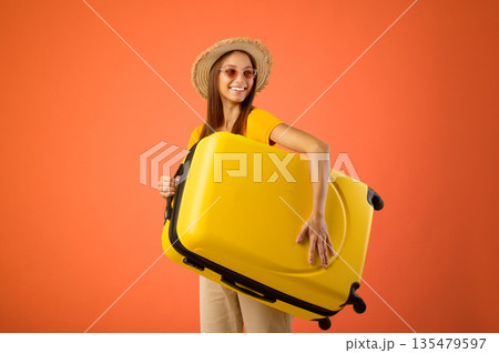 Pretty young woman in sunglasses and summer hat holding suitcase and smiling over yellow studio background, getting ready for vacation, getting amazing discounts and propositions, copy space 135479597