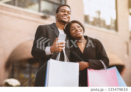 Great shopping on black friday and advertising of sale. Happy african american man and lady in coats with colored bags hug and look at something amazing, male holding smartphone near shop, copy space Great shopping on black friday and advertising of sale. Happy african american man and lady in coats with colored bags hug and look at something amazing, male holding smartphone near shop, copy space 135479610