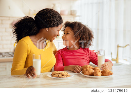 Happy Black Mother And Daughter Bonding Together While Having Snacks In Kitchen, Cheerful African American Mom And Her Little Child Sitting At Table Eating Cookies And Drinking Milk, Free Space 135479613