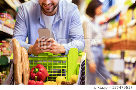 Closeup Of Unrecognizable Man Using Mobile Coupon App For Groceries Shopping Buying Food In Supermarket, Standing With Cart In Hypermarket. Smiling Guy Using Smartphone Purchasing Grocery In Shop 135479617