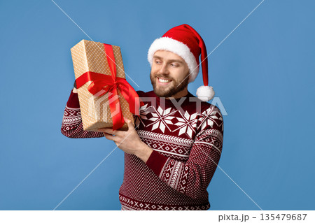 Handsome millennial guy in Christmas sweater and Santa hat holding wrapped gift box on blue studio background. Cheerful young man with New Year present anticipating winter holidays Handsome millennial guy in Christmas sweater and Santa hat holding wrapped gift box on blue studio background. Cheerful young man with New Year present anticipating winter holidays 135479687