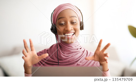 Attractive Muslim black woman in hijab wearing headphones, communicating online on laptop at home. Lovely female teacher in traditional headscarf giving remote lesson on webcam 135479709