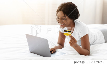 Online shopping from home, e-commerce concept. Happy young african american woman lying on bed at home and buying clothes or products online, paying with credit card via laptop, copy space Online shopping from home, e-commerce concept. Happy young african american woman lying on bed at home and buying clothes or products online, paying with credit card via laptop, copy space 135479745