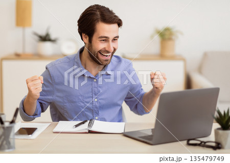 Joyful Guy At Laptop Computer Shaking Fists Gesturing Yes Celebrating Business Success Sitting In Modern Office. Great News And Victory Celebration Concept 135479749