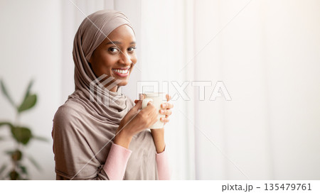 Black Muslim woman in traditional hijab drinking hot aromatic coffee near window at home, having peaceful weekend. Portrait of beautiful young lady in Arabic headscarf having warming beverage indoors 135479761