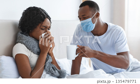 Black man in protective face mask taking care of his sick wife, giving hot drink, bedroom interior, empty space. African american woman with scarf around her neck having coronavirus, sneezing her noze 135479771