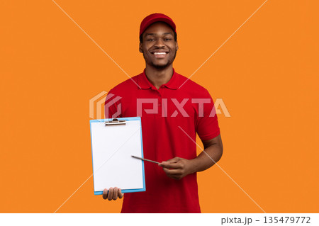 Parcel Receiving. Portrait Of Smiling African American Delivery Guy Holding Pen And Showing Clipboard With Blank Paper For Sign Here, Standing Isolated Over Orange Studio Background. Mock up Parcel Receiving. Portrait Of Smiling African American Delivery Guy Holding Pen And Showing Clipboard With Blank Paper For Sign Here, Standing Isolated Over Orange Studio Background. Mock up 135479772
