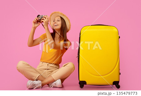 Happy girl taking photos on camera, sitting next to luggage over pink background Happy girl taking photos on camera, sitting next to luggage over pink background 135479773