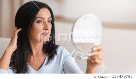 Portrait of beautiful middle-aged brunette lady holding mirror and looking at her reflection, touching her hair with hand. Confident adult woman thinking about skin care and body treatment Portrait of beautiful middle-aged brunette lady holding mirror and looking at her reflection, touching her hair with hand. Confident adult woman thinking about skin care and body treatment 135479783