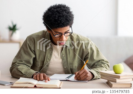 Remote learning. Black college student taking notes from textbook, studying at home during coronavirus outbreak. African American adolescent preparing for test or making homework indoors Remote learning. Black college student taking notes from textbook, studying at home during coronavirus outbreak. African American adolescent preparing for test or making homework indoors 135479785