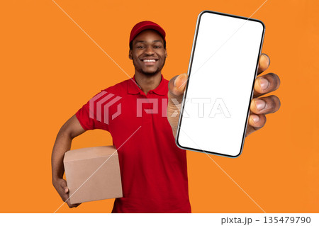 Easy Payment. Smiling african american male courier in red cap and uniform holding credit card and cardboard parcel box, carrying package, standing isolated over orange studio background Easy Payment. Smiling african american male courier in red cap and uniform holding credit card and cardboard parcel box, carrying package, standing isolated over orange studio background 135479790