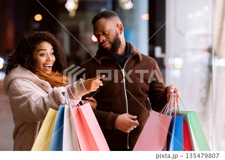 Omg, Let's Buy This. Excited african american woman pointing at shop window, walking with her husband near shopping mall in the evening. Seasonal Sales And Discount, Retail, Purchase 135479807