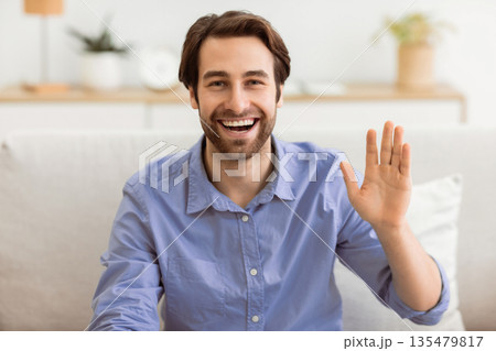 Hello. Happy Man Waving Hand Smiling To Camera Sitting On Couch At Home. Cheerful Guy Gesturing Hi Greeting Making Video Call Indoors. Modern Remote Communication Concept 135479817