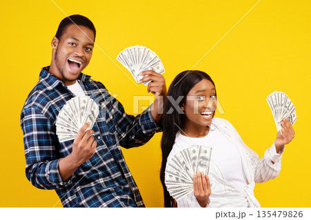 Get money, big win together and relationship. Happy millennial african american guy and woman dance and rejoice with a lot of dollars in hands, isolated on yellow background, studio shot, free space 135479826