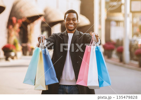 Beginning of sales season and Black Friday for shopaholic before holidays. Happy millennial african american man shows colored bags in camera near modern city mall after shopping, free space Beginning of sales season and Black Friday for shopaholic before holidays. Happy millennial african american man shows colored bags in camera near modern city mall after shopping, free space 135479828