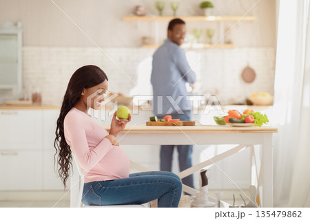 Beautiful pregnant woman with apple and her loving boyfriend cooking balanced meal at kitchen, copy space. Millennial expectant couple eating healthy at home. Perfect pregnancy diet concept 135479862