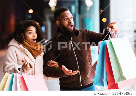 Wow, Look At It. Excited african american couple looking and pointing at shop window, surprised by offer, walking near shopping mall in the evening. Seasonal Sales And Discount, Purchase And Retail Wow, Look At It. Excited african american couple looking and pointing at shop window, surprised by offer, walking near shopping mall in the evening. Seasonal Sales And Discount, Purchase And Retail 135479863