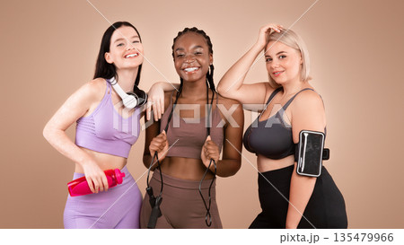 Three smiling multiracial women with different figure and skin type posing in sportswear over brown studio background, copy space Three smiling multiracial women with different figure and skin type posing in sportswear over brown studio background, copy space 135479966