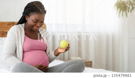 Pregnant Women Nutrition. Happy Black Expectant Lady Holding Green Apple While Relaxing On Bed At Home, Enjoying Healthy Eating, Smiling Awaiting Woman Tenderly Touching Her Belly, Copy Space 135479981