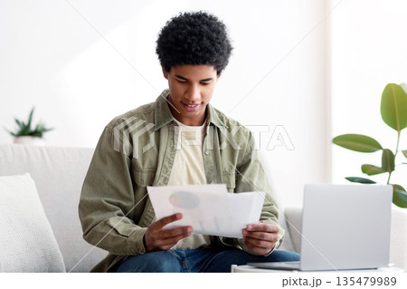 Happy black guy learning online from home, writing coursework, holding documents near laptop. African American teen student consulting with his teacher about educational project Happy black guy learning online from home, writing coursework, holding documents near laptop. African American teen student consulting with his teacher about educational project 135479989