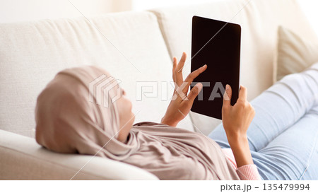African American woman in hijab with tablet computer lying on sofa at home, mockup for design on screen. Muslim lady studying or working online, watching movie or chatting with friend African American woman in hijab with tablet computer lying on sofa at home, mockup for design on screen. Muslim lady studying or working online, watching movie or chatting with friend 135479994