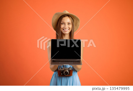 Happy young woman traveller holding laptop with blank screen over yellow studio background, website for travellers and tourists concept. Smiling lady tourist showing empty screen on her notebook 135479995