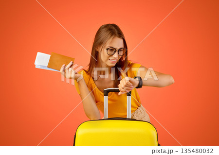 Worried young woman with passport and plane tickets looking at her watch, checking time, missed her flight. Sad lady traveller waiting for taxi, late to airport, yellow studio background, copy space 135480032