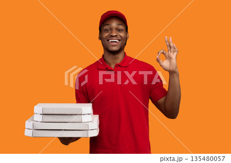 Fast Service And Best Food Delivery During Pandemic. Positive black courier wearing red uniform and cap holding pizza boxes from restaurant and showing okay sign gesture, isolated on orange background 135480057