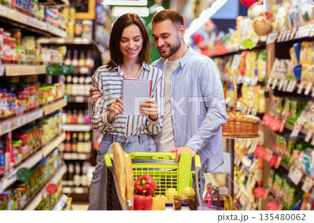 Young lovely couple with shopping cart full of healthy eco products looking into their grocery list at supermarket. Smiling man embaricng woman while buying products at mall together 135480062