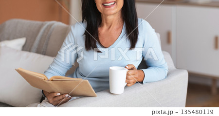 Leisure, Rest And Relax Concept. Portrait of smiling mature woman reading book and holding mug of coffee, sitting on the sofa in living room in her apartment, enjoying weekend and looking at camera Leisure, Rest And Relax Concept. Portrait of smiling mature woman reading book and holding mug of coffee, sitting on the sofa in living room in her apartment, enjoying weekend and looking at camera 135480119