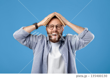 Furious young man putting book on his head and screaming on blue studio background. Bearded millennial guy tired of studying, cannot do difficult home assignment or meet deadline 135480130