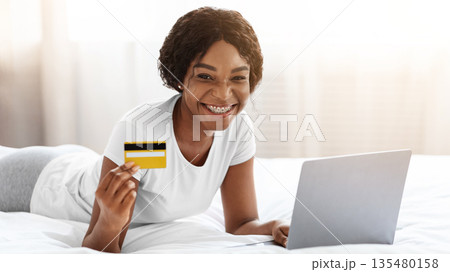 Shopping on internet from home, e-commerce concept. Cheerful young african american woman lying on bed at home and buying clothes or products online, paying with credit card via laptop, free space Shopping on internet from home, e-commerce concept. Cheerful young african american woman lying on bed at home and buying clothes or products online, paying with credit card via laptop, free space 135480158