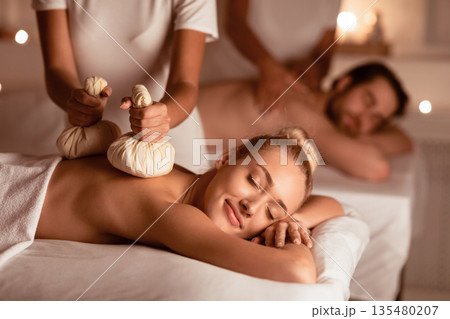 Couple Receiving Exotic Thai Herbal Massage With Aromatic Bags Enjoying And Relaxing Lying At Luxury Spa Resort During Honeymoon Vacation. Relaxation, Wellness, Beauty Procedure. Selective Focus Couple Receiving Exotic Thai Herbal Massage With Aromatic Bags Enjoying And Relaxing Lying At Luxury Spa Resort During Honeymoon Vacation. Relaxation, Wellness, Beauty Procedure. Selective Focus 135480207
