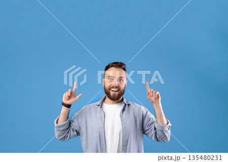 Excited bearded guy pointing up at blank space over blue studio background. Handsome millennial man having great offer, suggesting sale or discount, advertising your product Excited bearded guy pointing up at blank space over blue studio background. Handsome millennial man having great offer, suggesting sale or discount, advertising your product 135480231