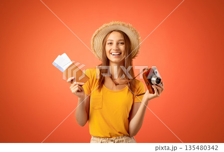 Smiling young woman in summer hat holding camera and passport with flight tickets in, yellow studio background 135480342