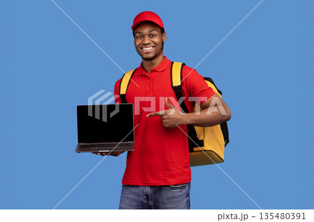 Order Here. Portrait of smiling black delivery man holding laptop and pointing at empty screen for mockup, wearing red uniform and yellow thermo backpack, standing on blue studio background, copyspace 135480391