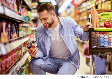 Consumption And Consumerism. Portrait Of Smiling Bearded Man With Shopping Cart In Market Buying Groceries Food Taking Products From Shelves In Store, Holding Glass Jar Of Sauce, Checking Label Consumption And Consumerism. Portrait Of Smiling Bearded Man With Shopping Cart In Market Buying Groceries Food Taking Products From Shelves In Store, Holding Glass Jar Of Sauce, Checking Label 135480449