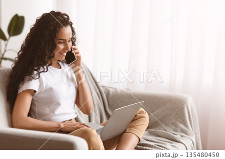 Happy young woman freelancer talking on mobile phone and using laptop, working from home during isolation, copy space. Curly lady having conversation on phone and typing on computer keyboard 135480450