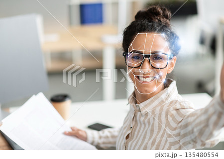 A person is using facial recognition technology while sitting at a desk in a contemporary office. They appear engaged and friendly, showcasing the ease of identification in today's work environment. 135480554